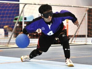 A goalball player wearing a blue jersey stands in front of a net preparing to throw the ball. 